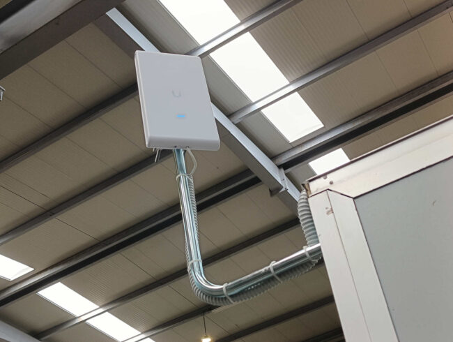 Instalación finalizada del punto de acceso Wi-Fi UniFi U6 Mesh Pro con cableado protegido en nave industrial de acero
